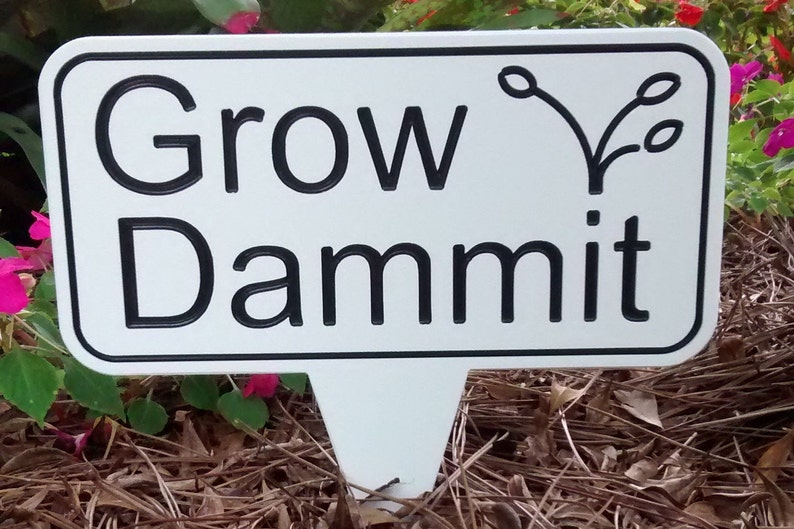 May include: A white garden stake with a black border and the words "Grow Dammit" in bold black letters. A stylized plant illustration is above the word "mit". The stake is in a garden setting with green foliage and pink flowers.