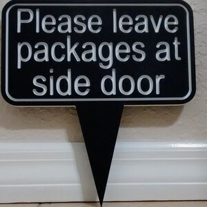 May include: Black sign with white text that reads "Please leave packages at side door". The sign is attached to a black stake.