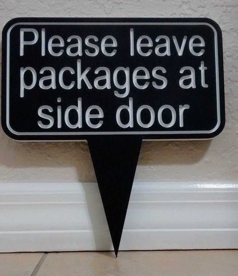 May include: Black sign with white text that reads "Please leave packages at side door". The sign is attached to a black stake.
