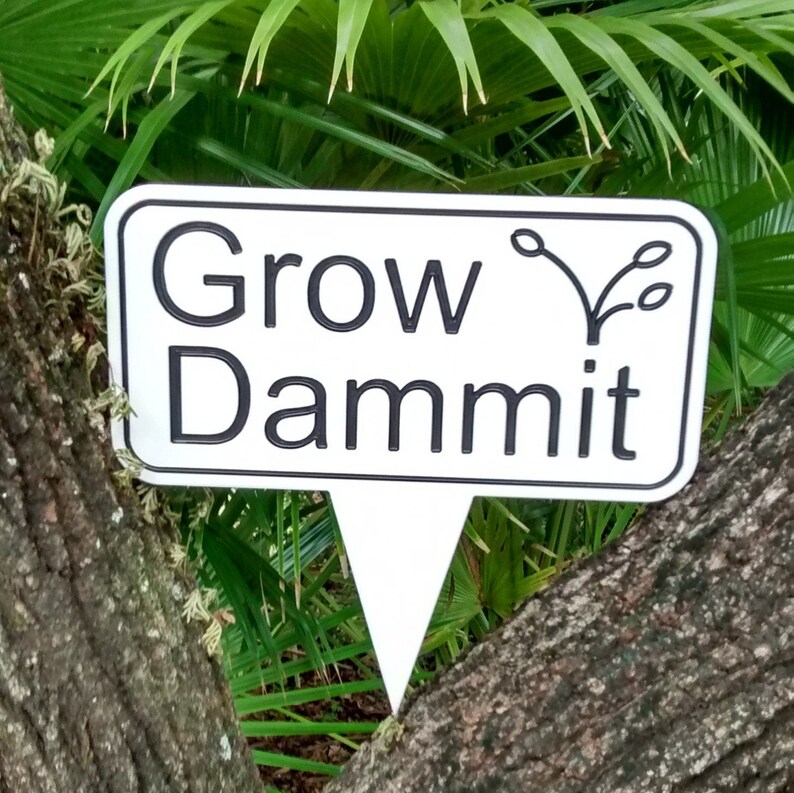 May include: A white rectangular garden stake with a black border and the words "Grow Dammit" in black, with a plant graphic. The stake is placed between two tree trunks, with green foliage in the background.