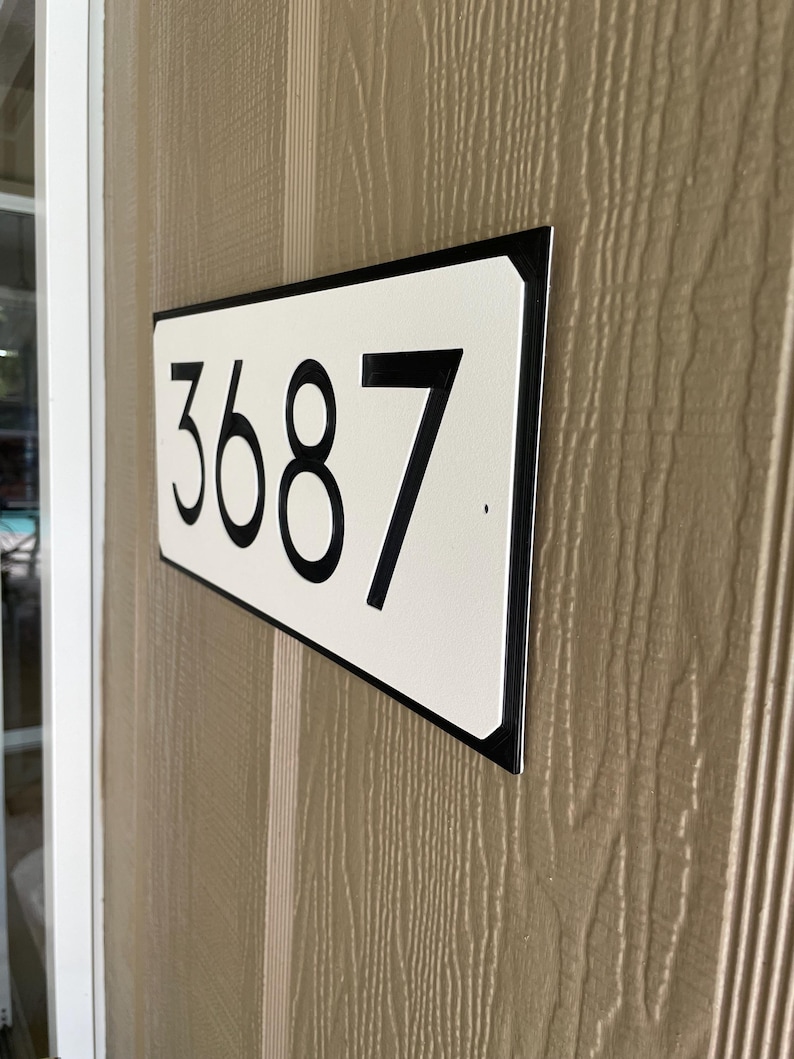 Pode incluir: Uma placa de n&uacute;mero de casa retangular branca com moldura preta e n&uacute;meros pretos. A placa diz "3687".
