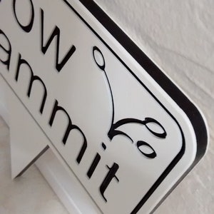 May include: A white sign with black lettering and a black border. The sign reads "row ammit" with a decorative flourish. The sign is mounted on a white support structure.