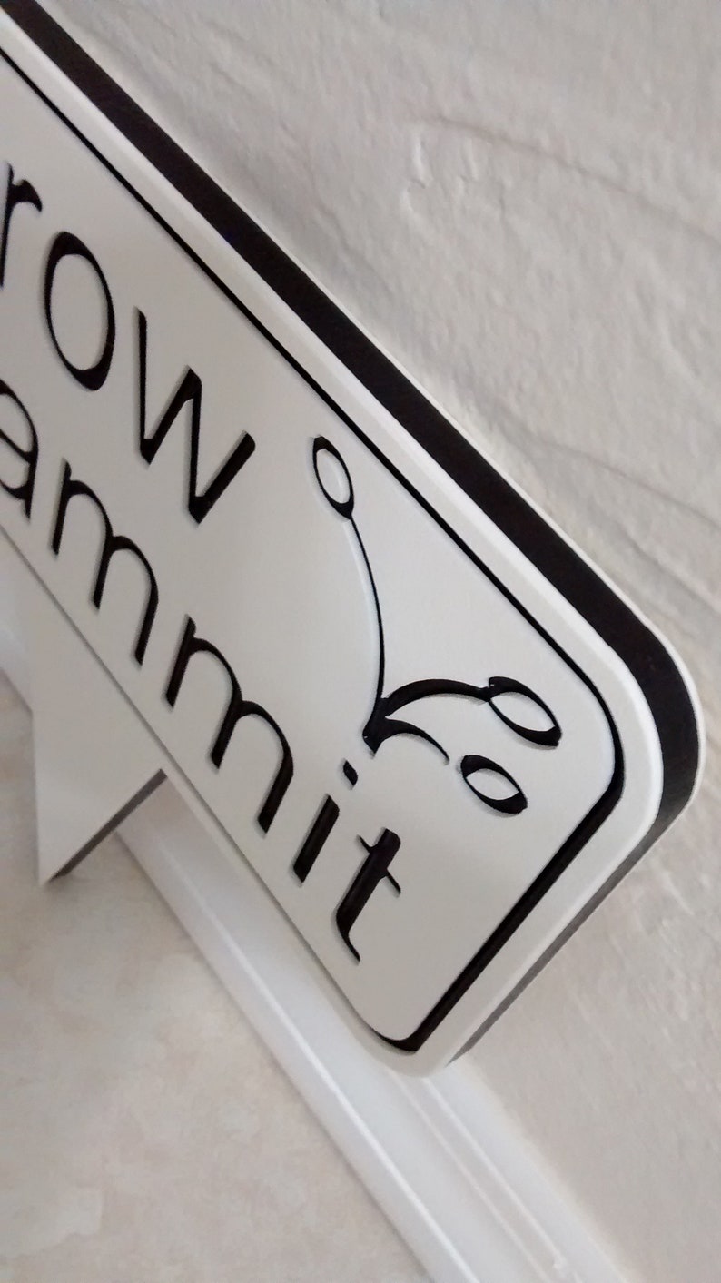 May include: A white sign with black lettering and a black border. The sign reads "row ammit" with a decorative flourish. The sign is mounted on a white support structure.