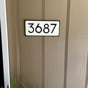 Pode incluir: Uma placa de n&uacute;mero de casa retangular branca com moldura preta e n&uacute;meros pretos. A placa diz "3687".