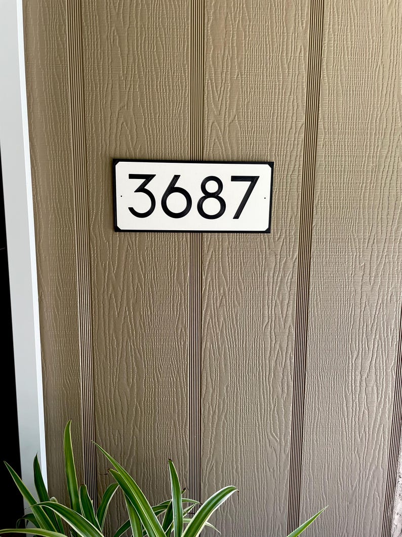 Pode incluir: Uma placa de n&uacute;mero de casa retangular branca com moldura preta e n&uacute;meros pretos. A placa diz "3687".