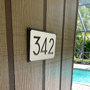 Pode incluir: Uma placa de n&uacute;mero de casa branca com moldura preta e o n&uacute;mero 342 em uma parede de madeira marrom.