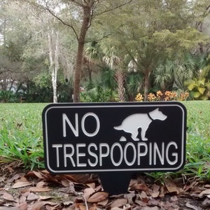 Op de afbeelding: Een zwart-wit bord met de tekst "NO TRESPOOPING" en een silhouet van een hond die poept. Het bord staat op een groen gazon met bruine bladeren.