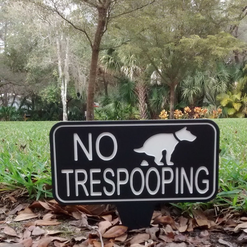 Op de afbeelding: Een zwart-wit bord met de tekst "NO TRESPOOPING" en een silhouet van een hond die poept. Het bord staat op een groen gazon met bruine bladeren.
