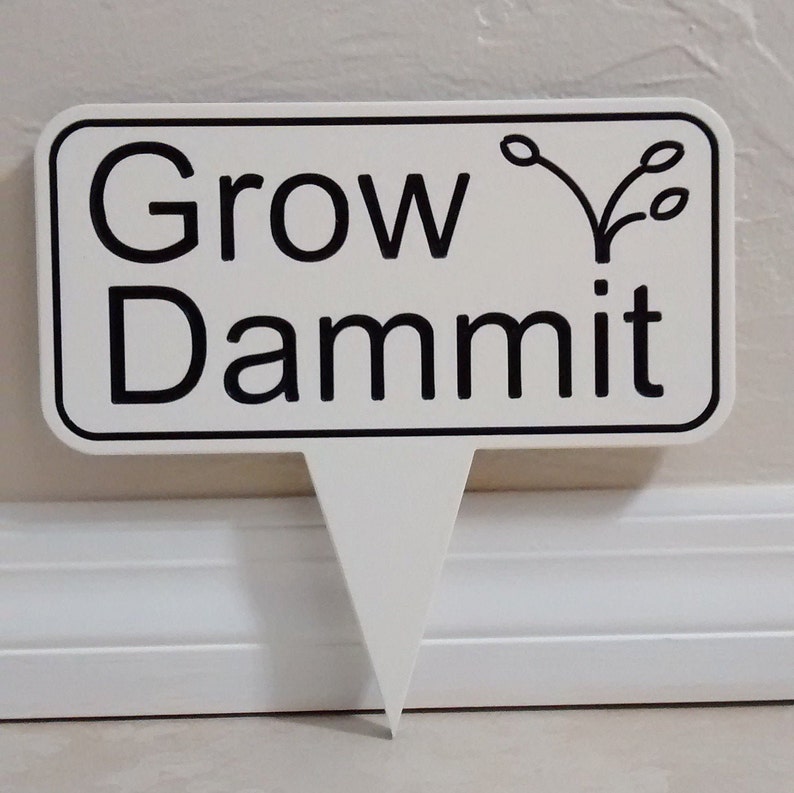 May include: A white rectangular garden stake with rounded corners and a black border. The text "Grow Dammit" is printed in black, with a small plant illustration above the word "Dammit".