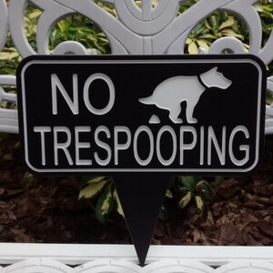 Op de afbeelding: Een zwart-wit bord met de tekst "NO TRESPOOPING" en een wit silhouet van een hond die poept. Het bord is gevormd als een puntige pijl.