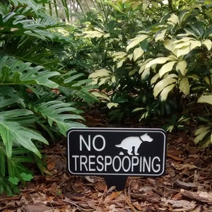 Op de afbeelding: Een zwart-wit bord met de tekst "NO TRESPOOPING" en een wit silhouet van een hond die poept. Het bord staat in een tuin met groene planten en bruine bladeren.