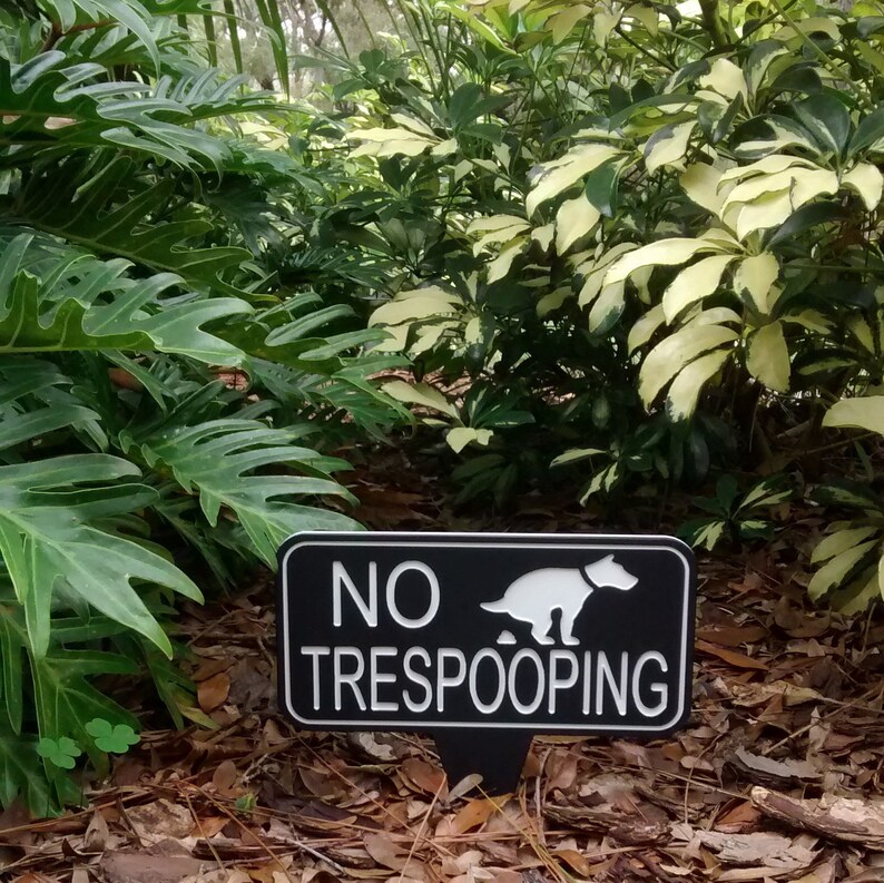 Op de afbeelding: Een zwart-wit bord met de tekst "NO TRESPOOPING" en een wit silhouet van een hond die poept. Het bord staat in een tuin met groene planten en bruine bladeren.