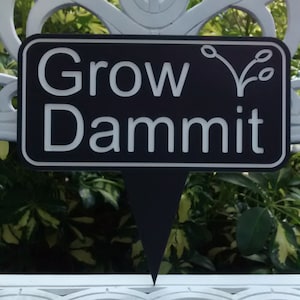 May include: A black and white garden stake with the words "Grow Dammit" in white lettering. The stake has a pointed bottom for easy insertion into the soil and a decorative plant icon.