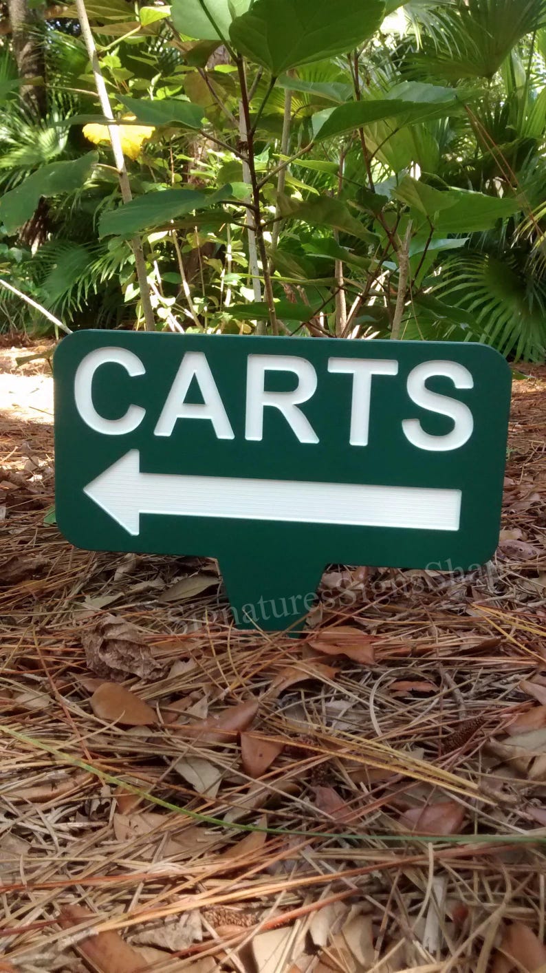 Puede incluir: Un letrero verde con letras blancas que dice "CARTS" con una flecha blanca apuntando hacia la izquierda. El letrero est&aacute; sobre un lecho de paja de pino.