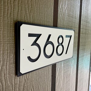 Pode incluir: Uma placa de n&uacute;mero de casa retangular branca com moldura preta e n&uacute;meros pretos. A placa diz "3687".