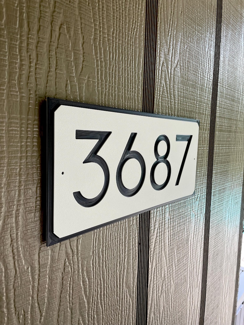 Pode incluir: Uma placa de n&uacute;mero de casa retangular branca com moldura preta e n&uacute;meros pretos. A placa diz "3687".