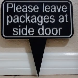 May include: Black sign with white text that reads "Please leave packages at side door". The sign is on a white wall and has a black stake.