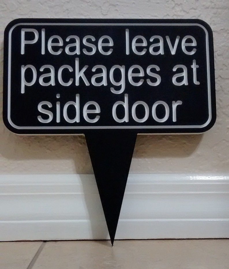 May include: Black sign with white text that reads "Please leave packages at side door". The sign is on a white wall and has a black stake.