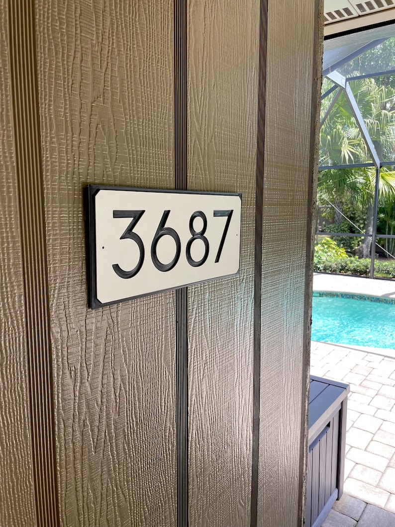 Pode incluir: Uma placa de n&uacute;mero de casa retangular branca com moldura preta e o n&uacute;mero 3687 impresso em preto.