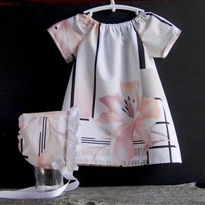 Puede incluir: Un vestido blanco con estampado floral negro y rosa y mangas cortas. Viene con un gorro a juego con ribete de encaje blanco.