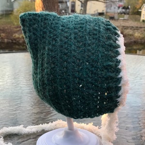 May include: A green crocheted baby bonnet with a white crocheted trim. The bonnet is displayed on a white stand.
