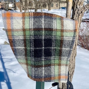 May include: A plaid fabric hood with orange, green, black and white colours. The hood is hanging on a wooden stick.