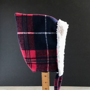 Puede incluir: Un gorro de bebé con capucha de cuadros rojos y negros con forro de sherpa blanco y lazos. El gorro mide aproximadamente 28 cm de alto.