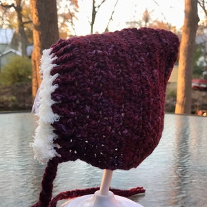 May include: A crocheted baby bonnet in burgundy and white. The bonnet has a white crocheted trim and a long burgundy tie.