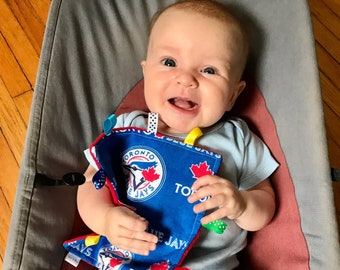 May include: A baby wearing a light blue onesie with a Toronto Blue Jays themed baby toy. The toy has a blue background with the team logo and the words "Toronto Blue Jays".