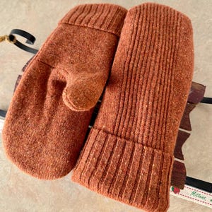 May include: A pair of rust-coloured knit mittens featuring a textured, ribbed cuff design. The mittens are crafted from a speckled, heathered yarn material, showcasing a consistent vertical knit pattern across the surface of both hand coverings.