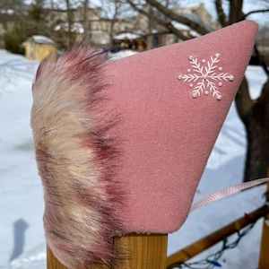 May include: A pink, pointed hat with a white snowflake design and faux fur trim. The hat is made of a soft material and has a ribbon attached. The hat is likely for a child.