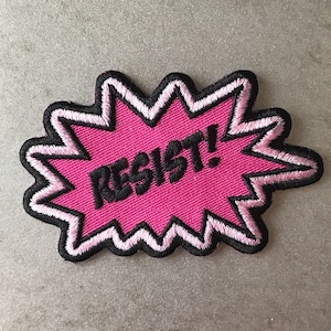 May include: Pink fabric patch with a white outline and black stitching. The patch is shaped like a starburst and says "RESIST!" in black letters.