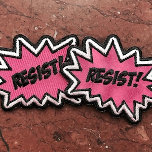 May include: Two pink fabric patches with a white outline and black stitching. The patches are shaped like a starburst and say "RESIST!" in black lettering.