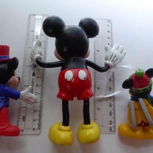 K&ouml;nnte beinhalten: Drei Mickey-Mouse-Figuren. Die mittlere Figur hat ausgestreckte Arme, rote Shorts und gelbe Schuhe. Die anderen beiden Figuren sind in einem blauen Anzug und einem Winteroutfit gekleidet. Ein Lineal befindet sich hinter den Figuren.