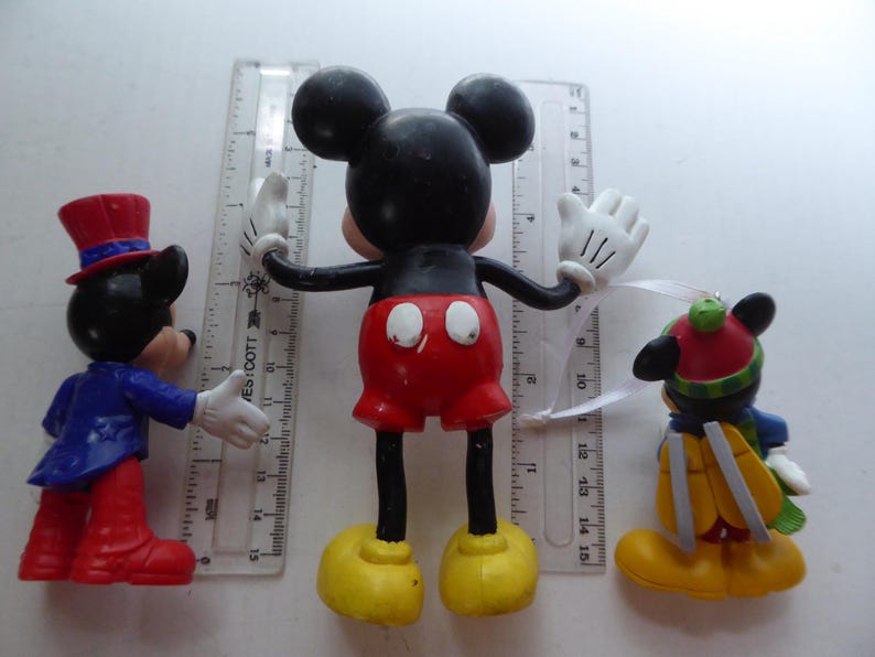 K&ouml;nnte beinhalten: Drei Mickey-Mouse-Figuren. Die mittlere Figur hat ausgestreckte Arme, rote Shorts und gelbe Schuhe. Die anderen beiden Figuren sind in einem blauen Anzug und einem Winteroutfit gekleidet. Ein Lineal befindet sich hinter den Figuren.