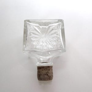 May include: Clear glass bottle with a square top and a starburst design. The bottle has a cork stopper. The bottle is empty and sits on a white surface.