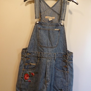 May include: Pale blue denim dungaree shorts with adjustable straps and pockets. Features embroidered red roses on the front. The shorts are hanging on a black hanger. The brand name is visible on the pockets.