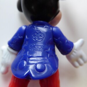 K&ouml;nnte beinhalten: Eine Mickey Mouse-Figur, von hinten gesehen, tr&auml;gt eine blaue Jacke mit dem Text "EPCOT CENTER". Die Figur hat wei&szlig;e Handschuhe und eine rote Hose. Die Jacke hat ein Logo auf dem R&uuml;cken.
