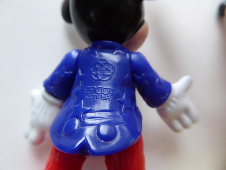 K&ouml;nnte beinhalten: Eine Mickey Mouse-Figur, von hinten gesehen, tr&auml;gt eine blaue Jacke mit dem Text "EPCOT CENTER". Die Figur hat wei&szlig;e Handschuhe und eine rote Hose. Die Jacke hat ein Logo auf dem R&uuml;cken.