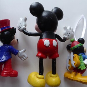 K&ouml;nnte beinhalten: Drei Mickey-Mouse-Figuren. Eine tr&auml;gt eine blaue Jacke und rote Hosen, eine andere hat die Arme in einer klassischen Pose ausgestreckt, und die dritte ist mit Schlittschuhen f&uuml;r den Winter gekleidet. Alle haben schwarze Ohren, wei&szlig;e Handschuhe und gelbe Schuhe.