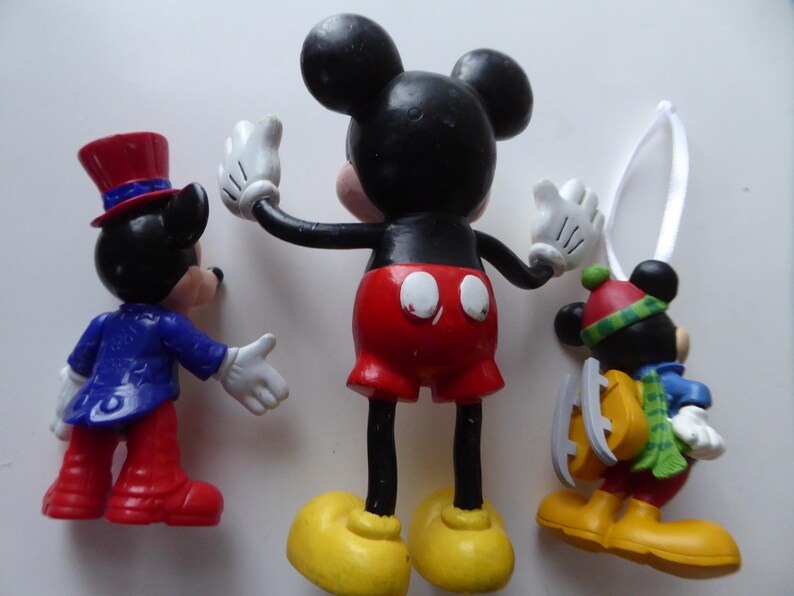 K&ouml;nnte beinhalten: Drei Mickey-Mouse-Figuren. Eine tr&auml;gt eine blaue Jacke und rote Hosen, eine andere hat die Arme in einer klassischen Pose ausgestreckt, und die dritte ist mit Schlittschuhen f&uuml;r den Winter gekleidet. Alle haben schwarze Ohren, wei&szlig;e Handschuhe und gelbe Schuhe.