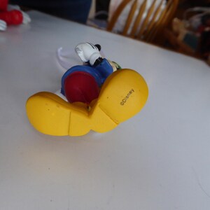 K&ouml;nnte beinhalten: Eine Mickey Mouse-Figur mit gelben Schuhen, rotem Unterteil, blauer Jacke und wei&szlig;en Handschuhen. Die Figur liegt auf der Seite, mit dem Wort "Disney" auf der Schuhsohle.