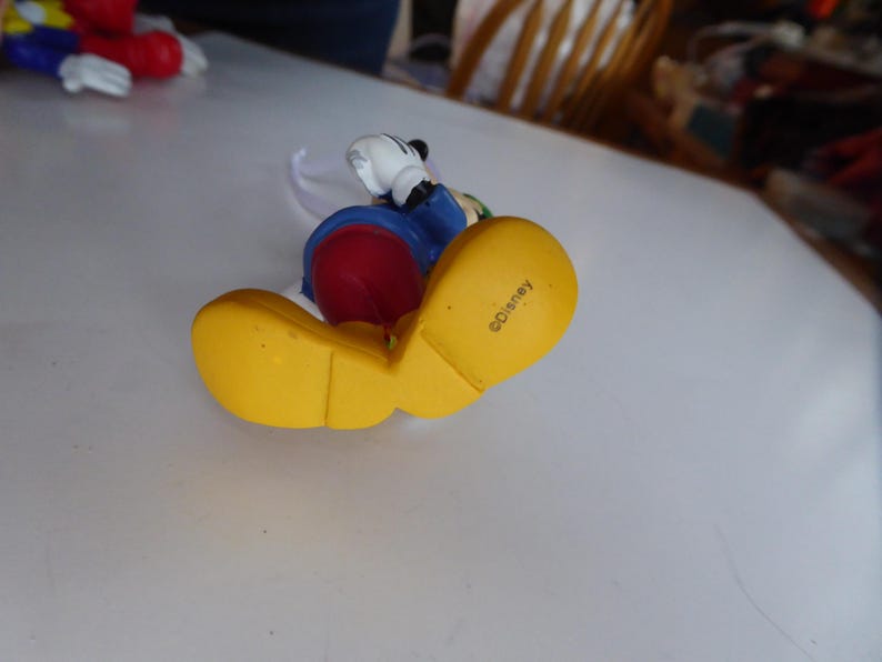 K&ouml;nnte beinhalten: Eine Mickey Mouse-Figur mit gelben Schuhen, rotem Unterteil, blauer Jacke und wei&szlig;en Handschuhen. Die Figur liegt auf der Seite, mit dem Wort "Disney" auf der Schuhsohle.