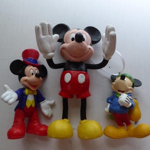 K&ouml;nnte beinhalten: Drei Mickey Mouse-Figuren. Die mittlere Figur hat die Arme erhoben und tr&auml;gt rote Shorts und gelbe Schuhe. Die Figur links tr&auml;gt einen Zylinder und eine blaue Jacke. Die Figur rechts tr&auml;gt eine blaue Jacke und eine gr&uuml;ne M&uuml;tze.