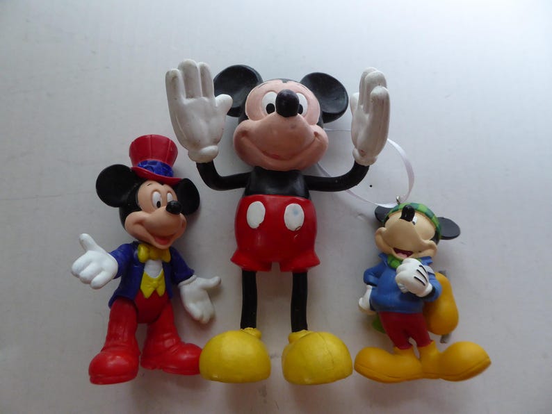 K&ouml;nnte beinhalten: Drei Mickey Mouse-Figuren. Die mittlere Figur hat die Arme erhoben und tr&auml;gt rote Shorts und gelbe Schuhe. Die Figur links tr&auml;gt einen Zylinder und eine blaue Jacke. Die Figur rechts tr&auml;gt eine blaue Jacke und eine gr&uuml;ne M&uuml;tze.