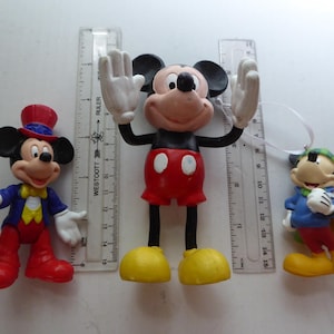K&ouml;nnte beinhalten: Drei Mickey-Mouse-Figuren. Ein Mickey tr&auml;gt eine blaue Jacke, eine rote Hose und einen Zylinder. Ein anderer Mickey hat die Arme erhoben und tr&auml;gt rote Shorts und gelbe Schuhe. Der dritte Mickey tr&auml;gt eine blaue Jacke und eine gr&uuml;ne M&uuml;tze.