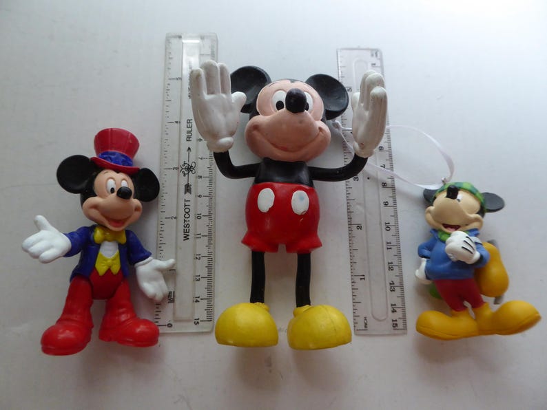 K&ouml;nnte beinhalten: Drei Mickey-Mouse-Figuren. Ein Mickey tr&auml;gt eine blaue Jacke, eine rote Hose und einen Zylinder. Ein anderer Mickey hat die Arme erhoben und tr&auml;gt rote Shorts und gelbe Schuhe. Der dritte Mickey tr&auml;gt eine blaue Jacke und eine gr&uuml;ne M&uuml;tze.