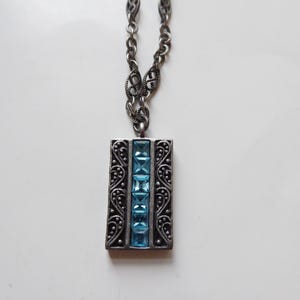 May include: A silver necklace with a rectangular pendant featuring a row of blue gemstones set in a decorative silver frame.