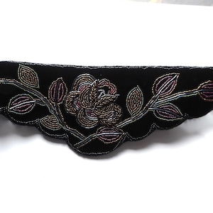 Op de afbeelding: Zwarte fluwelen riem met een bloemenontwerp van kralen. De riem heeft een centrale roos en bladeren in goud-, paars- en bruintinten. De randen van de riem hebben een geschulpte vormgeving. Een decoratief accessoire.