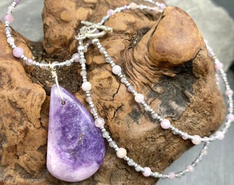 May include: A delicate necklace with a large, teardrop-shaped purple amethyst pendant. The necklace is made with small, light pink and silver beads.